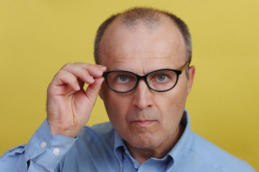 Handsome man 45-55 years old in blue shirt on yellow background holding his glasses with his hand.