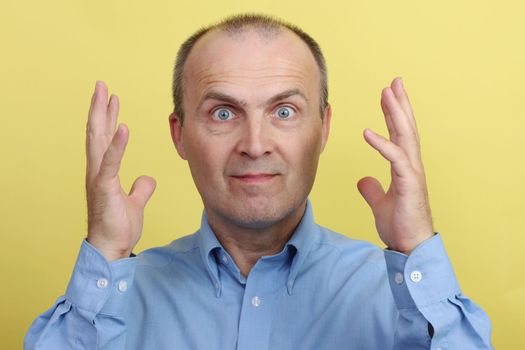 A handsome elderly man in a blue shirt on a yellow background gesticulates emotionally with his hands.