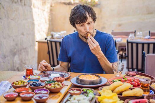 Man eating turkish breakfast. Turkish breakfast table. Pastries. Vegetables. Olives. Cheeses, fried eggs. Jams, tea in copper pot and tulip glasses. Wide composition.