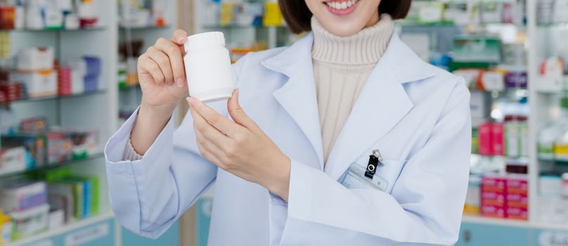 Closeup young affable pharmacist show qualified medicine pill bottle mockup product for copyspace at pharmacy. Druggist working with her diary job at drugstore, medicine box on shelf at background