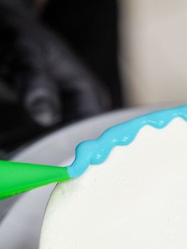 pastry chef cake designer decorating turquoise blue white frosted cheesecake in kitchen with piping bag