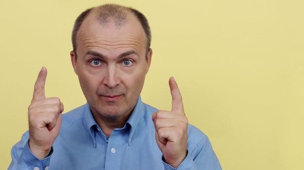 A man 45-55 years old in a blue shirt on a yellow background raised two hands with an index finger.