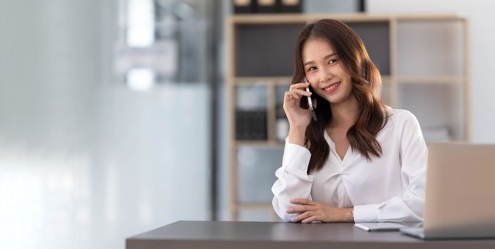 Young Asian businesswoman sit with her laptop computer happily talking on the phone with her customer explaining the detail in the office...
