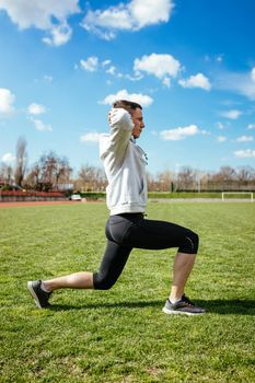 Active young man stretching and doing one leg crouching exercises in the public place on empty stadium. 