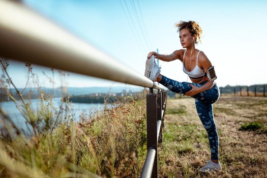 Young urban runner woman with headphones doing stretching exercises on the fence after jogging. She is doing workout on the meadow with a view on the river.