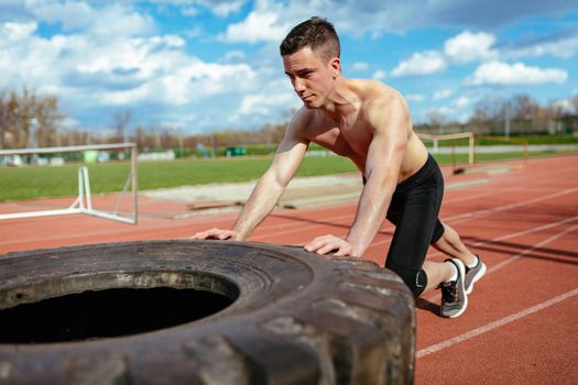Young muscular man pushing up a huge tire for training muscles at the tartan track on the stadium.