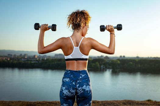 Rear view of a young fitness woman doing workout with dumbbells by the river in a sunset.