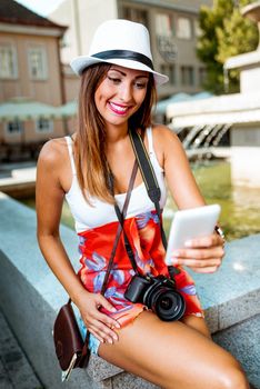 Beautiful young woman on vacation taking selfie with her smart phone in the city square next to the fountain.