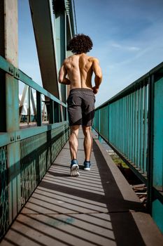 Rear view of a fit muscular young male runner with naked torso, sprinting at great speed outdoors on the bridge.