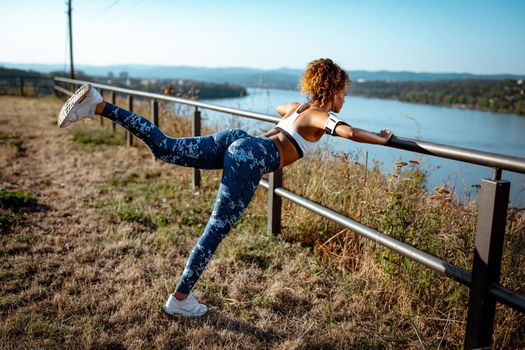 Young urban runner woman with headphones doing stretching exercises on the fence after jogging. She is doing workout on the meadow with a view on the river.