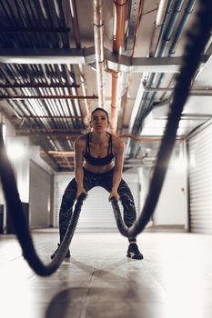 Muscular young woman exercising with ropes at the garage gym. Selective focus.