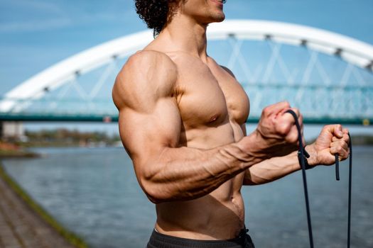 Close up on a naked torso of strong bodybuilder with six pack, perfect ABS, shoulders, biceps, triceps and chest, who is doing strong fit body training with elastic rubber band, near the river. 