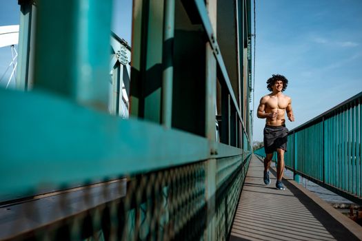 Fit muscular young male runner with naked torso, sprinting at great speed outdoors on the bridge.