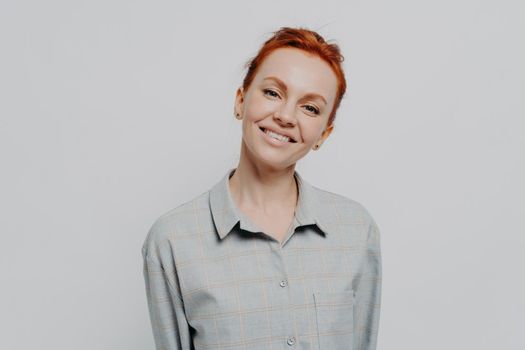 Cheerful happy beautiful red haired woman with beaming smile posing isolated on grey studio background, dressed in casual checkered shirt, expressing positivity and happiness. Face expressions concept