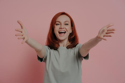 Free cuddles. Friendly happy ginger woman reaching hands forward and stretching arms for hug and smiling cheerfully while standing against pink wall, dressed in casual t-shirt