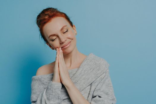 Happy calm redhead grateful woman holding hands in prayer gesture with closed eyes and tilted head, waiting for miracle with smile on face isolated on white blue studio background. Faith and hope