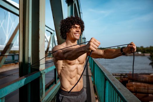 Bodybuilder with naked torso, is doing strong fit body training with elastic rubber band, on the river bridge.  
