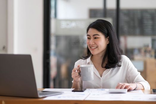 Charming Asian businesswoman sitting in the office with a digital laptop computer. Excited Asian businesswoman raising hands to congratulate while working in a modern office,..