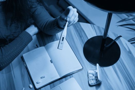 sick young woman with pills and thermometer on a table working on a laptop from home on quarantine isolation. Online work, stay home concept