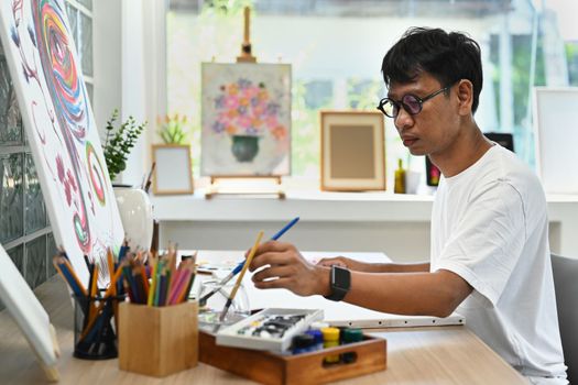Asian man artist sitting in art studio and painting picture on canvas with oil paints. Art and leisure activity concept.