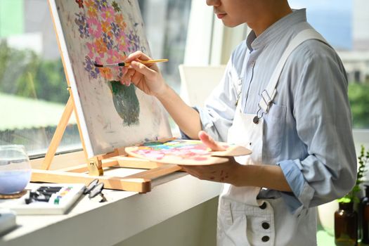Cropped shot of artist with palette and brush painting on canvas near window in modern workshop.
