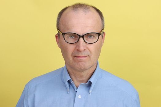 A man in a blue shirt and glasses poses against a yellow background and looks at the camera.