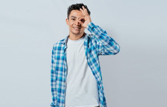 Happy young man making ok gesture with hand looking through fingers. Friendly man making ok gesture looking through fingers. Teenage guy making ok gesture looking through fingers