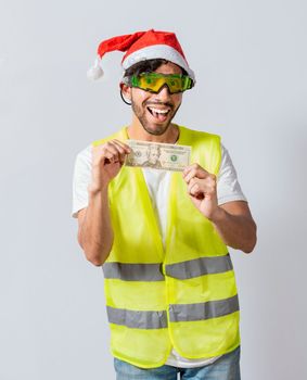 Happy builder engineer with christmas hat holding one dollar bill isolated. Happy christmas engineer showing one dollar bill, Happy engineer in vest and christmas hat showing twenty dollars