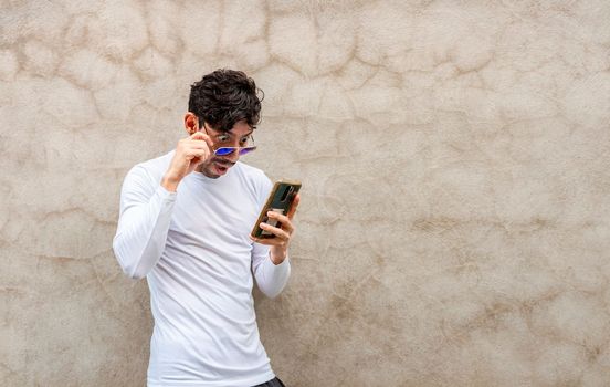 Surprised young Latin looking at his cell phone leaning against a wall. Handsome guy in glasses amazed with his cell phone leaning against a wall, Latin man looking phone leaning against a wall