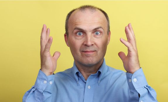 A handsome elderly man in a blue shirt on a yellow background gesticulates emotionally with his hands.