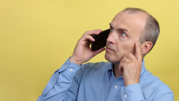 Portrait of a man in profile with a smartphone in his hand and the other hand touches his face. Elderly man with a phone in a blue shirt on a yellow background.