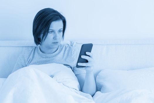 Woman using smartphone as she sits in bed covered with a duvet