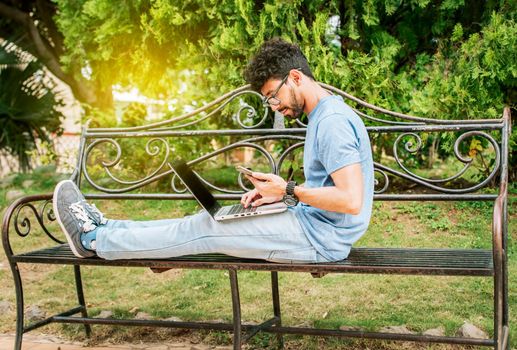 Freelancer man sitting in a park using laptop and cellphone. Man in a park working online with laptop. Relaxed man working with laptop outdoor, Young man in a park with laptop and cell phone