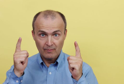 A man 45-55 years old in a blue shirt on a yellow background raised two hands with an index finger.