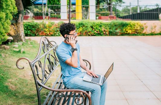 Young man sitting in a park working with his laptop and cell phone. Man in a park working online with laptop and calling on cellphone. Freelancer man working with laptop while calling on the phone