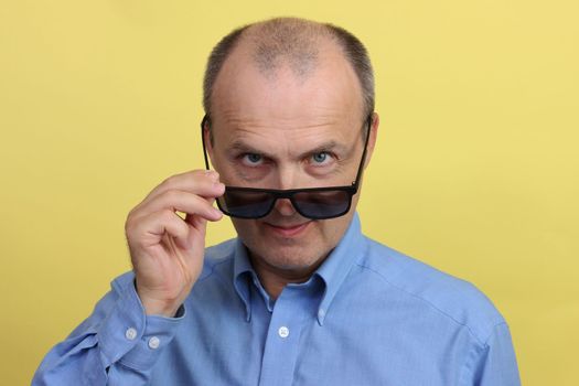 Close-up portrait of a positive man in a blue shirt holding black glasses in his hand looking at the camera.