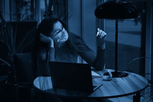 sick young woman with pills and thermometer on a table working on a laptop from home on quarantine isolation. Online work, stay home concept