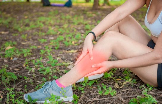 Athlete girl sitting on the grass with leg pain. Sore sporty girl sitting on the grass rubbing her leg. Outdoor athlete woman leg pains concept. Athlete woman rubbing her leg sitting on the grass