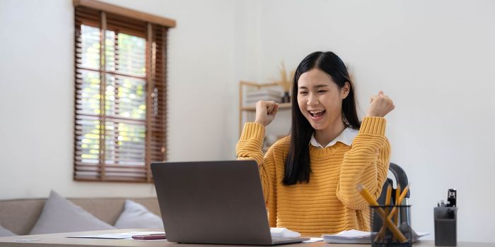 Excited happy woman looking at the laptop computer screen, celebrating an online win, overjoyed young asian female screaming with joy at home.