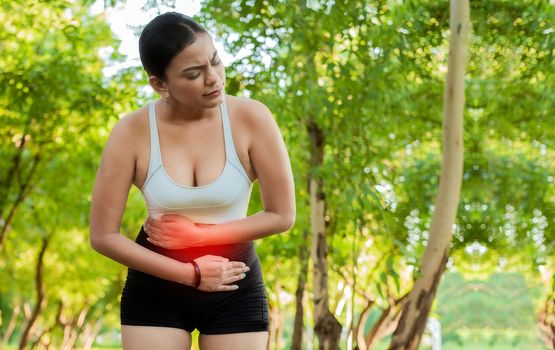 Woman in sportswear with stomach ache in a park. Running woman with digestive problems. Sport girl with stomach ache outdoors with copy space. Fatigued young woman with stomach ache