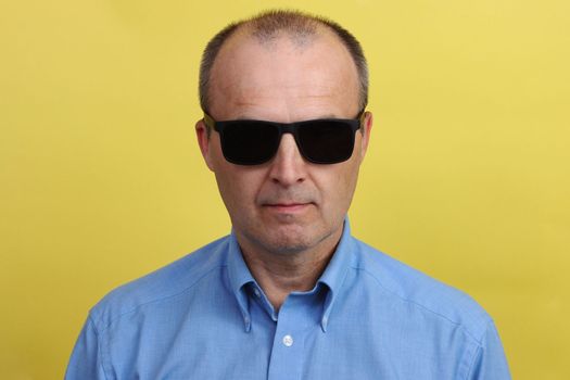 Handsome man 55 years old in a blue shirt in black sunglasses on a yellow background.