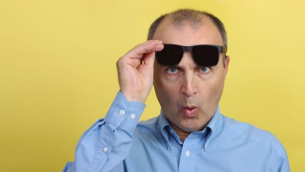 A man with glasses on a yellow background. Handsome surprised man in a blue shirt on a yellow background holds black glasses in his hands and looks at the camera.