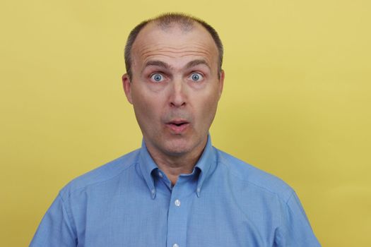 Portrait of a surprised handsome man 45-55 years old in a blue shirt looks at the camera.A man in a blue shirt on a yellow background.