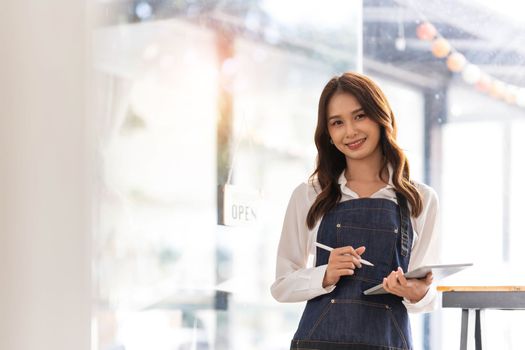 Beautiful asian young barista woman in apron holding tablet and standing in front of the door of cafe with open sign board. Business owner startup concept...