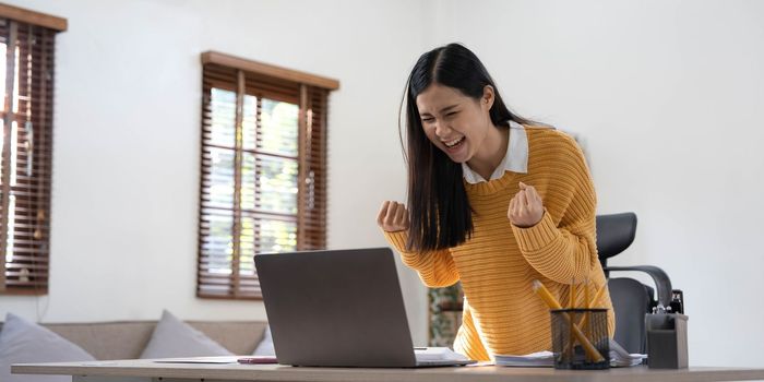 Excited happy woman looking at the laptop computer screen, celebrating an online win, overjoyed young asian female screaming with joy at home.