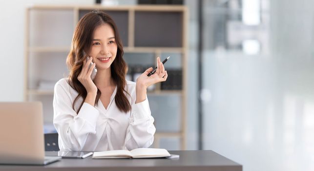 Young Asian businesswoman sit with her laptop computer happily talking on the phone with her customer explaining the detail in the office...