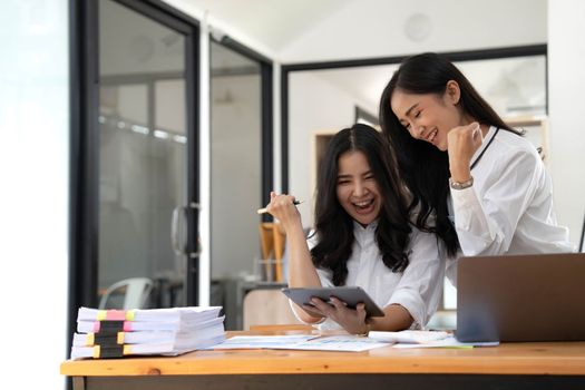 Two young Asian businesswomen show joyful expression of success at work smiling happily with a laptop computer in a modern office..