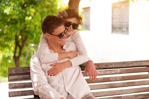 Smiling beautiful woman and her handsome boyfriend. Woman embrace summer day. Happy cheerful couple in sunglasses walking old city park. Couple posing on the street near green tree