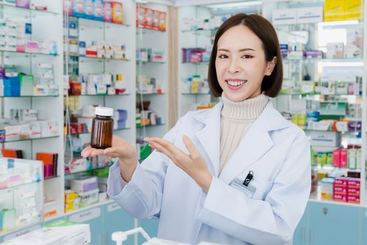 Portrait of young affable pharmacist show qualified medicine pill bottle mockup product for copyspace at pharmacy. Druggist working with her diary job at drugstore, medicine box on shelf at background