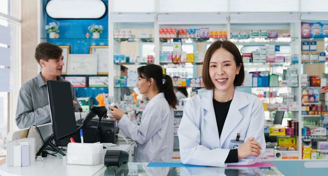 Portrait of affable asian pharmacist and qualified pharmaceutical, medicine pill bottle on shelf in background at pharmacy. Concept of pharmacist working on cashier talking to customer in drugstore.